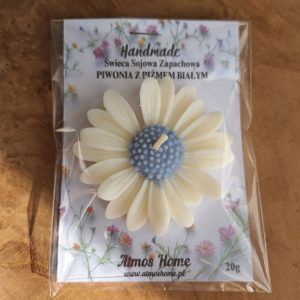 Zdjęcie przedstawia ręcznie robioną świecę sojową w kształcie piwonii. Świeca jest koloru kremowego z niebieskimi akcentami. W tle widać delikatne kwiaty. Na świecy znajduje się etykieta z napisem "Handmade" oraz nazwą zapachu "Piwonia z białym piżmem". Produkt AtmosHome.pl.