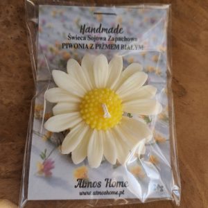 Zdjęcie przedstawia ręcznie robioną świecę sojową w kształcie piwonii. Świeca jest koloru kremowego z żółtymi akcentami. W tle widać delikatne kwiaty. Na świecy znajduje się etykieta z napisem "Handmade" oraz nazwą zapachu "Piwonia z białym piżmem". Produkt AtmosHome.pl.